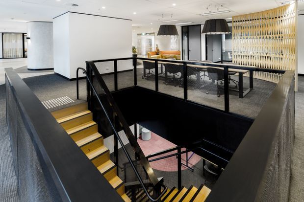 Contemporary commercial interior viewed from an upper-level landing, centred on a dramatic stair void with black metal mesh balustrades and contrasting yellow timber stair treads. The open-plan office includes carpeted floors, white walls and ceilings, meeting tables, pendant lights, and a decorative vertical timber screen.