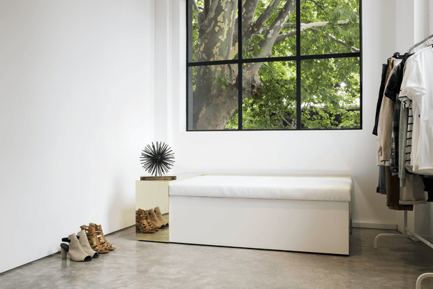 Bright minimalist dressing and wardrobe area with white walls, a large black-framed grid window overlooking leafy trees, a long white upholstered storage bench, freestanding clothing rack, shoes, and a sculptural sunburst decor piece. The room has a polished concrete floor and a clean, refined aesthetic.