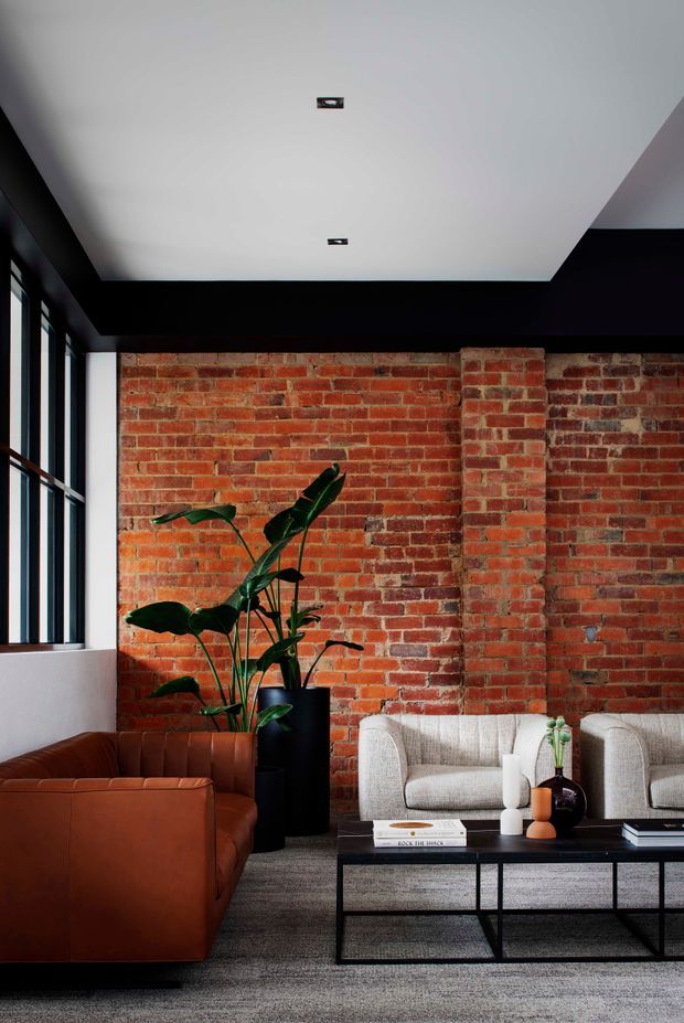 A sophisticated living room with exposed red brick walls, black structural framing, tall industrial-style windows, and a white recessed ceiling. The space contains a cognac leather sofa, two light upholstered armchairs, a black metal coffee table, and a tall indoor plant. The overall design combines industrial architectural elements with refined contemporary furnishings.