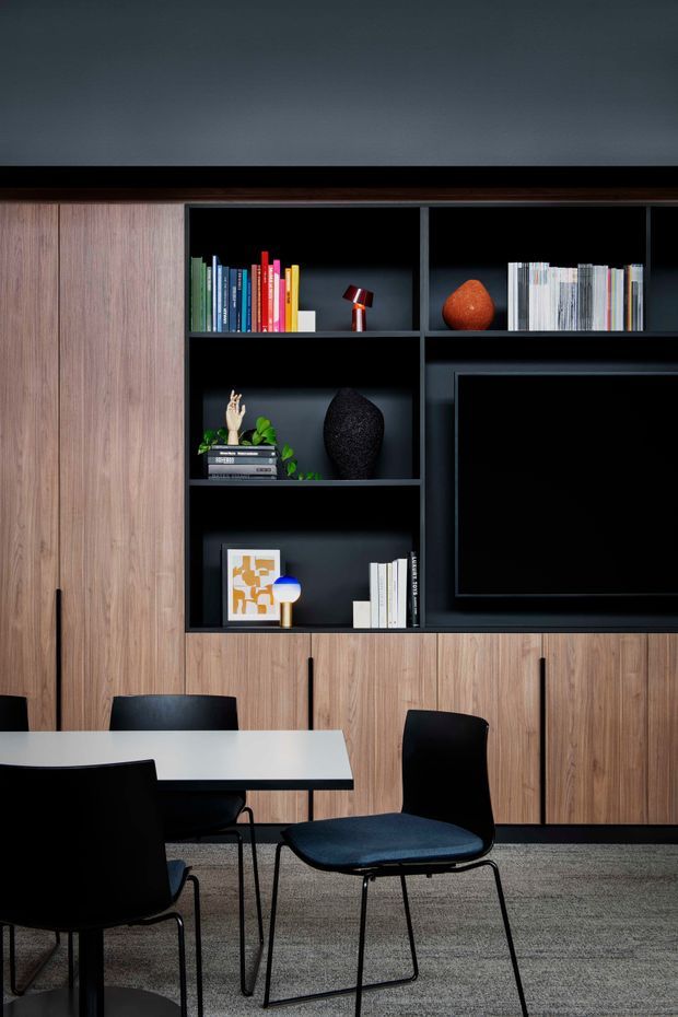 A dark, refined dining or living area centered on a full-height timber-look built-in wall unit. The joinery combines warm vertical wood cabinetry with matte black open shelving and an integrated television, displaying books, ceramics, plants, and small decorative objects. A white rectangular table and black contemporary dining chairs occupy the foreground, with grey carpet underfoot and a dark painted wall above the joinery.