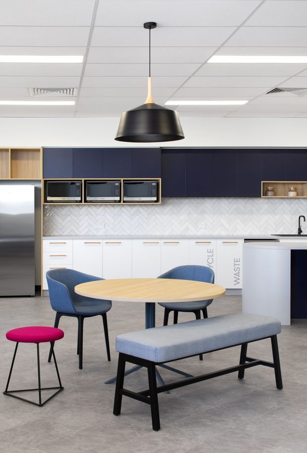 A contemporary commercial-style kitchen and dining/breakout area with dark navy cabinetry, white cabinetry, a white herringbone tile splashback, integrated appliances, and a stainless-steel refrigerator. A light timber oval table is surrounded by blue upholstered chairs and a long grey upholstered bench, with a bold pink stool in the foreground. A large black pendant light hangs beneath a suspended grid ceiling with fluorescent lighting.
