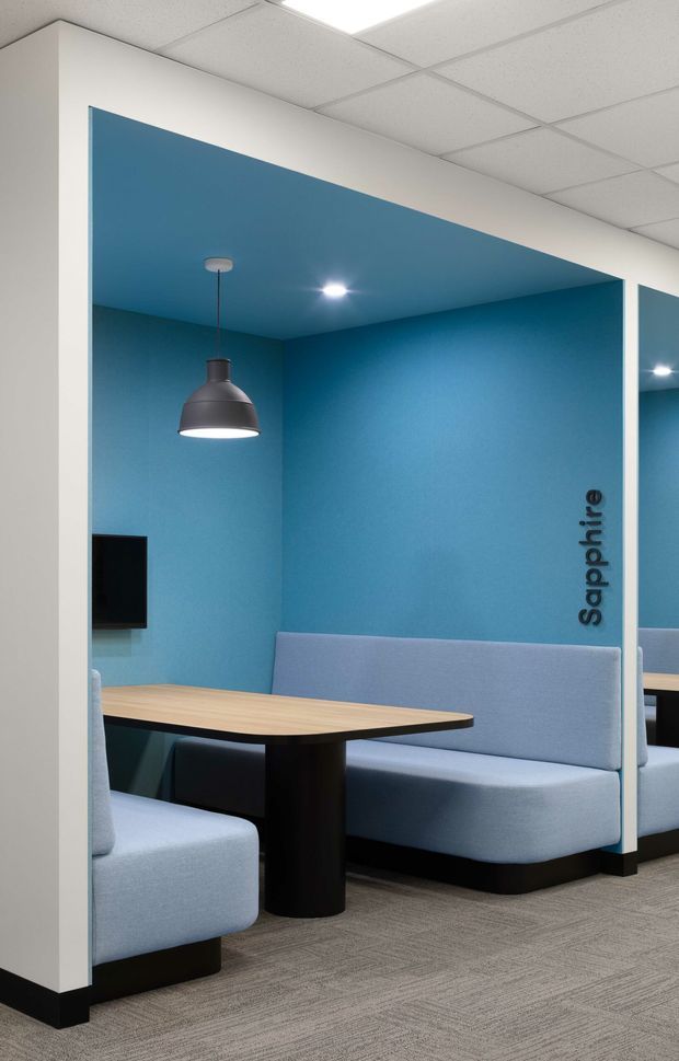 A modern commercial breakout or meeting booth with bright blue walls and ceiling, pale upholstered booth seating, a light timber-look table with black pedestal base, pendant and recessed lighting, and grey carpet. White structural framing creates an open cubicle layout, with a wall-mounted screen and the word “Sapphire” visible on the wall.