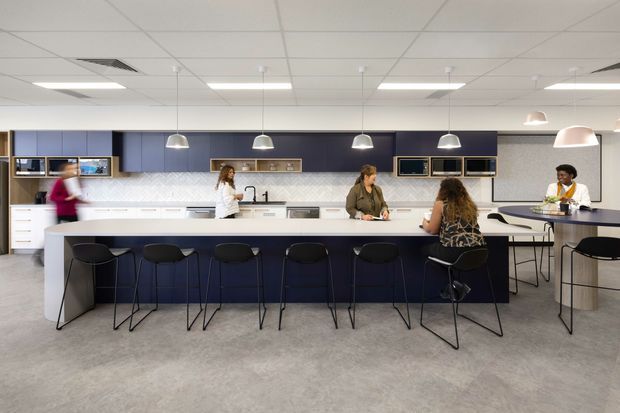 Contemporary communal kitchen and dining space with a long white island, dark blue fixed cabinetry, white geometric tile splashback, integrated appliances, and numerous black bar stools. Pendant lights hang above the island, while several people use the kitchen and raised dining counter. The room has a large open-plan commercial or workplace-breakout character with a suspended acoustic tile ceiling and grey flooring.