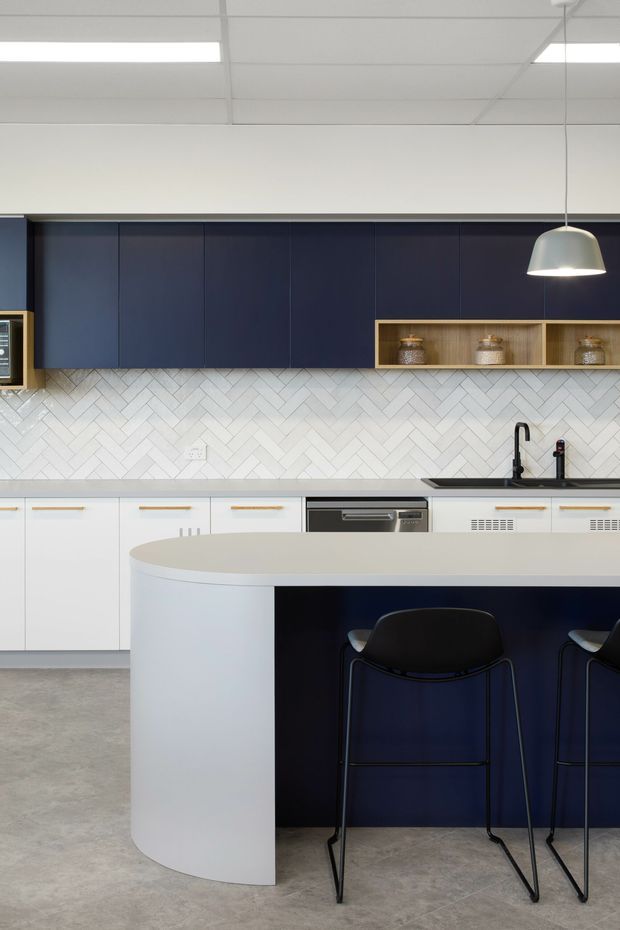 Contemporary kitchen with a large curved island, deep navy cabinetry, white lower cabinets, and a white herringbone tile splashback. The island includes a dark navy panel and integrated seating for multiple stools. A black sink tap, stainless-steel dishwasher, open timber display shelving, and minimalist pendant lighting complete the clean, modern fit-out.