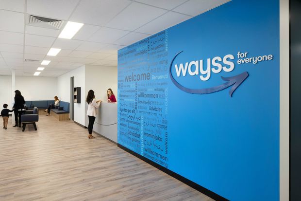 Bright contemporary commercial reception and waiting area with a large blue branded feature wall, white reception desk, light timber-look flooring, white walls, and suspended acoustic ceiling tiles with integrated lighting. People are present in the lobby, which opens into a small seating and waiting area.