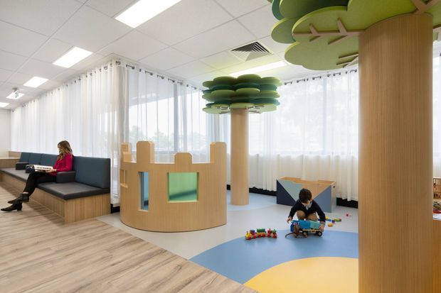 Bright contemporary children's play and waiting area with playful tree-shaped columns, a rounded timber play enclosure, colourful floor zones, and built-in seating beside floor-to-ceiling sheer curtains. A child is playing with toys while an adult sits on a dark upholstered bench. The space has pale timber-look flooring, white acoustic ceiling tiles, and abundant natural light.