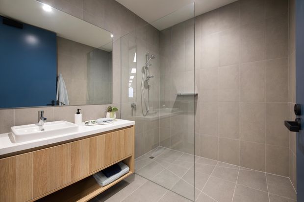 Contemporary bathroom with a floating timber vanity, white basin, large frameless mirror, and a glass-enclosed walk-in shower. Large-format grey tiles cover the walls and floor, with a deep blue door adding contrast.