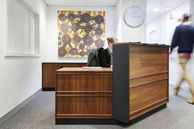 A bright contemporary reception area with a substantial timber reception desk and matching low cabinetry, set against white walls and grey carpet. A colourful geometric artwork and wall clock provide visual accents, while a corridor with people walking is visible to the right.