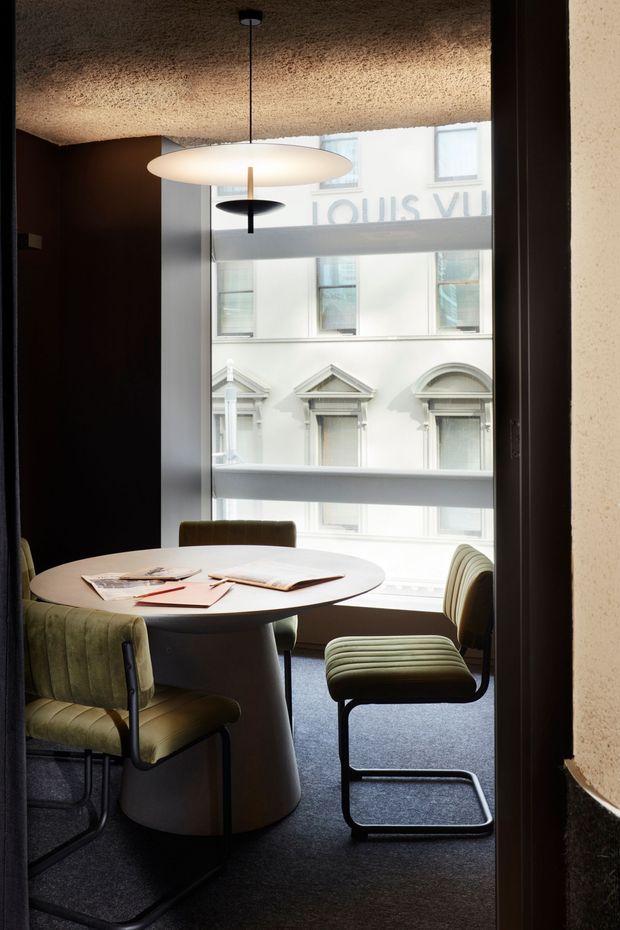 A compact, refined dining area beside a large street-facing window. A round white pedestal table is surrounded by upholstered green dining chairs and a matching low bench, with magazines on the tabletop. The room has dark walls, dark carpet, a textured acoustic ceiling, and a suspended oval pendant light. Historic decorative windows of an adjoining building are visible outside.