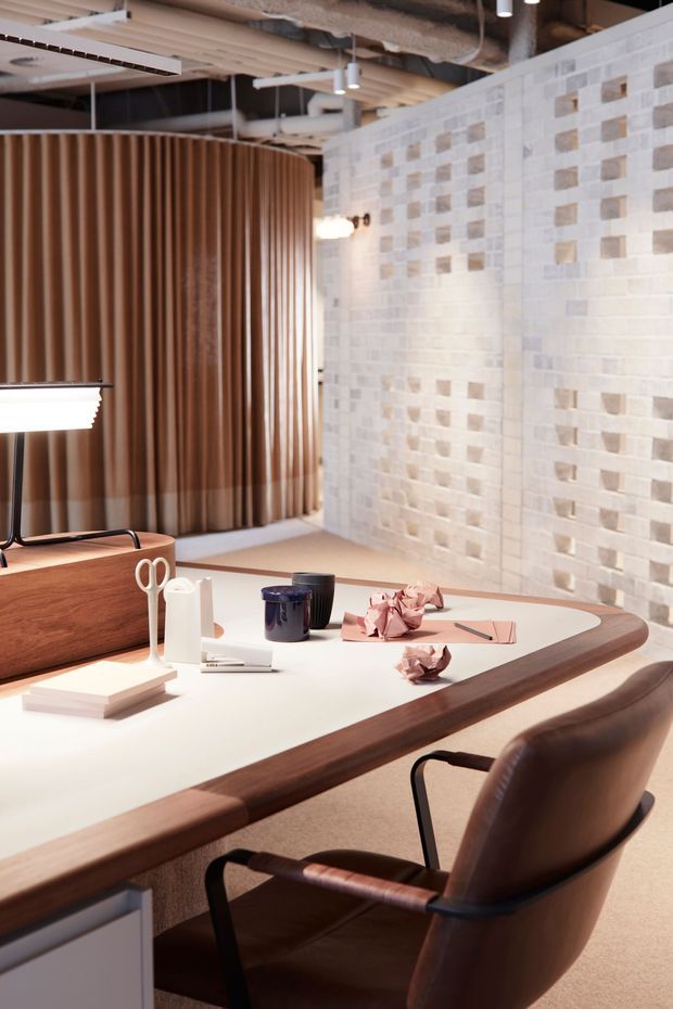 A refined office or meeting workspace with a large rounded rectangular desk in dark timber and a pale solid-surface top, paired with a sculptural brown leather armchair. The room features a curved vertical timber slat partition, a white and beige patterned masonry feature wall, exposed ceiling services and ducts, suspended linear and pendant lighting, and carefully arranged stationery and desk accessories. The overall aesthetic is warm, contemporary, and subtly industrial.