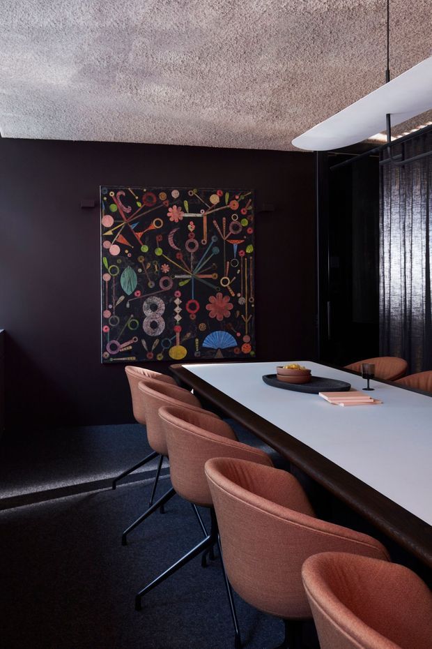 A moody, sophisticated dining room with a long white rectangular dining table and dark timber edge, surrounded by upholstered terracotta-toned dining chairs. The room has near-black walls, dark carpet, a textured pale ceiling, sheer dark window coverings, and a sculptural white linear pendant light. A colourful abstract geometric artwork provides a bold focal point on the wall.