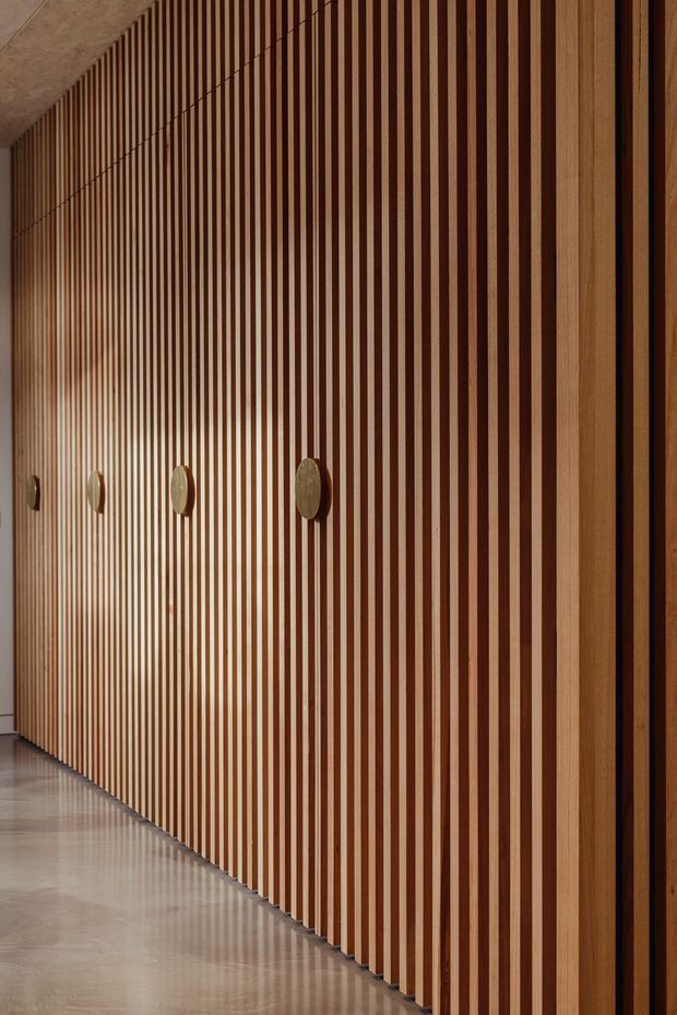 A close-up view of a long built-in wall of vertically ribbed timber cabinetry or panelling, with alternating warm wood slats and dark recessed gaps. Several round metal or stone pulls are spaced along the concealed doors. The installation runs through a refined corridor or entry area, with a smooth polished concrete or stone floor and a pale ceiling visible at the edges. The image has a warm, minimalist, highly detailed architectural interior aesthetic.