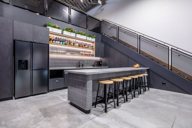A spacious, dark-toned industrial-style kitchen and home bar beneath a staircase. The room features charcoal cabinetry, a large black refrigerator, a tiled island with integrated sink, open bottle shelving, under-cabinet lighting, and a long row of timber-seated black bar stools. Exposed ceiling services, metal mesh stair balustrading, and a polished concrete floor reinforce the industrial character.
