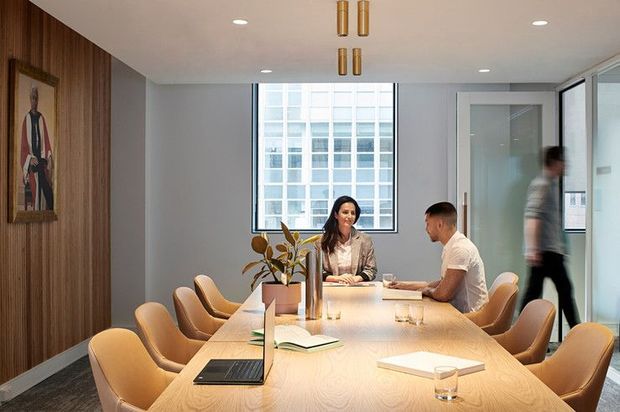 A contemporary dining or meeting room with a long light-wood table surrounded by tan upholstered chairs. The room features a full-height dark timber feature wall with framed artwork, pale painted walls, recessed ceiling lights, brass pendant lights, and large windows overlooking a neighbouring building. A glass door opens to an adjacent space.