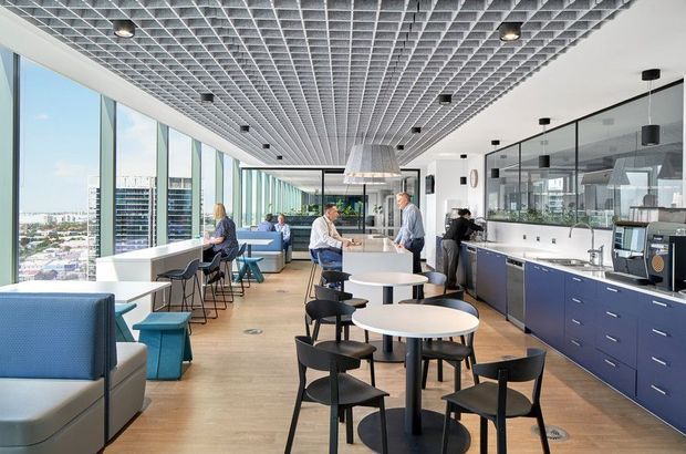 Bright contemporary commercial office breakout and café area with floor-to-ceiling glazing, a large communal kitchen, informal booth seating, bar stools, round dining tables, and a distinctive suspended grid ceiling. The space overlooks an urban setting and is furnished in a clean blue, white, black, and timber palette.