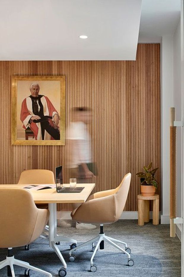 A refined office or meeting room with a rectangular light-wood table, upholstered tan swivel chairs, and a grey carpet. The room features a full-height vertical timber wall lining, white ceiling with recessed lighting, a framed portrait, and a small potted plant on a timber side table. A blurred figure and glass partition or doorway are visible at the right.