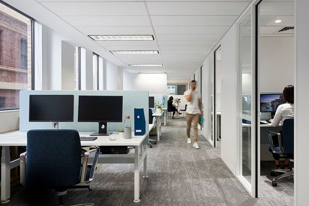 Bright contemporary open-plan office interior with rows of white workstations, dual computer monitors, ergonomic task chairs, glazed private offices along one side, large windows, suspended acoustic ceiling tiles, recessed fluorescent lighting, and grey patterned carpet flooring. The space has a clean, functional commercial workplace design.