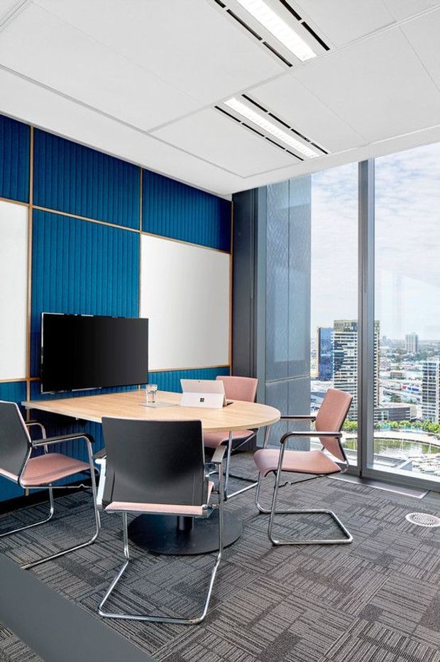 A contemporary commercial meeting room with a rounded light-wood conference table, black and blush-pink cantilever chairs, wall-mounted display, and a laptop. The room features bold blue vertical wall panelling with brass-toned trim, grey carpet tiles, a white suspended ceiling with linear recessed lighting, and full-height glazing overlooking a dense city skyline.