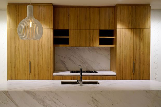 A contemporary kitchen viewed from the living side, featuring full-height warm timber cabinetry, a large white marble-look island with integrated sink and tap, and a matching marble splashback behind a built-in cooktop. Open shelving and a sculptural pendant light add visual interest, while white patterned wall surfaces are visible at either side.