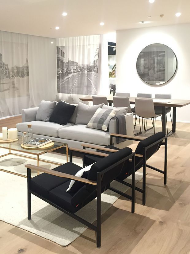 Bright open-plan living and dining area with a contemporary monochromatic fit-out. A light grey sofa and black-framed lounge chairs surround a glass-topped coffee table, while a timber dining table with upholstered chairs sits behind. Pale timber flooring, white walls, recessed ceiling lights, sheer curtains, large black-and-white wall artwork, and a circular wall mirror create a clean, refined interior.