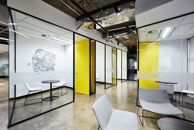 Open-plan commercial office interior with multiple glass-partitioned meeting rooms and workspaces. The space features black-framed glazing, bright yellow doors and frosted graphic film, exposed services and insulation at the ceiling, polished concrete flooring, and minimalist grey seating and tables.