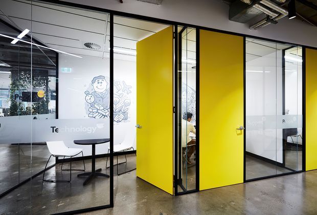 Contemporary open-plan office interior with exposed services, polished concrete flooring, extensive black-framed glass partitions, and vivid yellow meeting-room doors. White meeting tables and chairs sit within the glazed workspaces, with playful wall graphics and a strong industrial-commercial character.