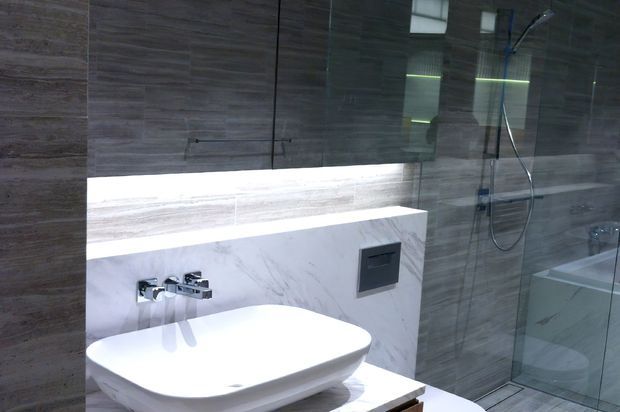 Modern bathroom featuring a white vessel basin on a white marble-look vanity, wall-mounted chrome tapware, dark wood-look wall cabinetry, large grey-brown stone-look wall tiles, and a frameless glass shower enclosure. Integrated lighting beneath the cabinetry creates a refined, contemporary atmosphere.