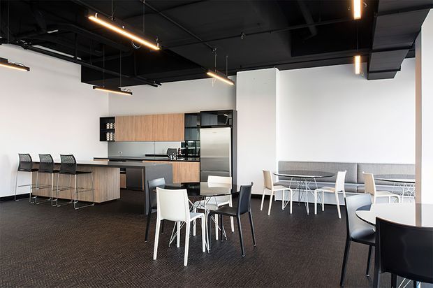An open-plan contemporary commercial breakout area featuring a compact kitchenette with timber cabinetry, dark benchtops, stainless-steel appliances, bar seating, and several dining or meeting tables. The space has a black exposed services ceiling with suspended linear lighting, white walls, dark carpet flooring, and a clean modern workplace aesthetic.