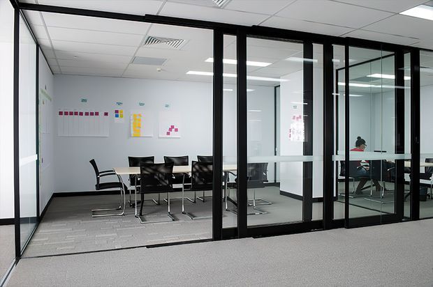 A contemporary commercial office interior with a glazed meeting room and adjacent open workspace. The meeting room has black-framed glass partitions, a rectangular conference table, black office chairs, white walls, grey carpet tiles, and a suspended acoustic ceiling with recessed fluorescent lighting. Additional desks and an occupied workstation are visible through the glazed partitions.