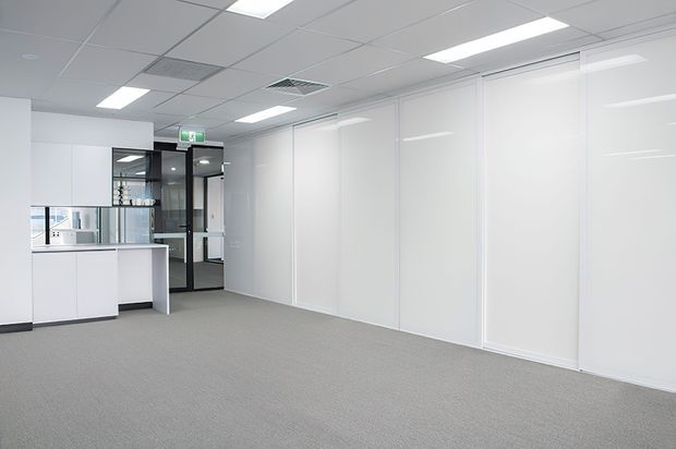 Bright, empty contemporary office interior with grey carpet, white full-height partition walls and built-in cabinetry, a suspended acoustic ceiling with recessed fluorescent lighting, and a glazed black-framed doorway leading to an adjacent office area.