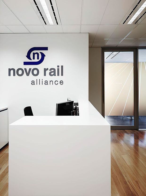 A minimalist commercial reception area featuring a large white reception desk, a white branded wall with the “novo rail alliance” logo, computer equipment, glass-partitioned offices, a suspended grid ceiling with linear fluorescent lighting, and timber flooring.