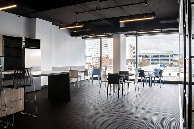 An open-plan contemporary commercial interior with a compact kitchenette or service counter on the left and multiple dining or meeting tables arranged beside expansive floor-to-ceiling windows. The space features a dark exposed ceiling with suspended linear lighting, white walls, black built-in joinery, dark patterned flooring, and views of an urban setting outside.