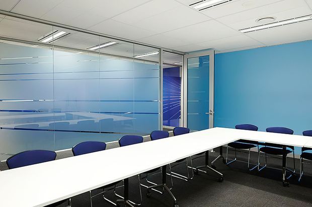 A contemporary commercial meeting or conference room with a long white boardroom table surrounded by blue office chairs. The room has bright blue painted walls, extensive frosted and clear glass partitioning with blue privacy bands, a blue glass door, grey carpet flooring, and a suspended acoustic ceiling with linear fluorescent lighting.
