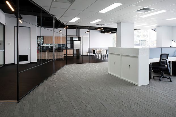 Contemporary open-plan commercial office interior with grey carpet tile flooring, white ceiling grid, glass-partitioned meeting and collaboration rooms, timber-toned cabinetry, and workstation desks with office chairs. Black-framed glazed partitions and linear pendant lighting create a refined, modern workplace environment.