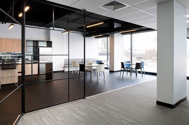 Open-plan contemporary commercial office interior with black-framed glazed meeting rooms, a compact kitchenette, shared tables and chairs, expansive windows, exposed black ceiling services, and carpeted circulation areas.
