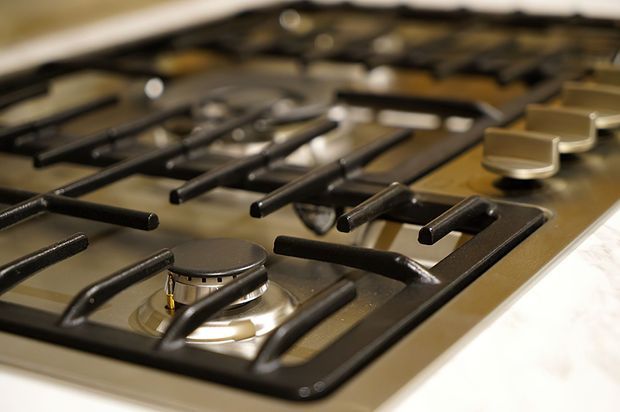 Close-up of a built-in stainless steel gas cooktop with multiple burners, black cast-iron pan supports, and metallic control knobs. The appliance is shown as part of a kitchen benchtop, with a shallow depth of field emphasizing the foreground burner.