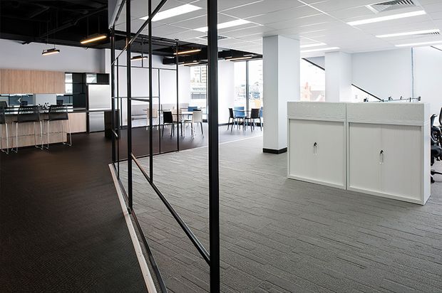 A bright contemporary open-plan office interior with grey carpet flooring, white partitions and columns, black-framed glazed meeting-room screens, and a kitchenette or breakout area with timber cabinetry at the rear. Exposed black services and linear pendant lighting contrast with the suspended white acoustic ceiling. Workstations and a small meeting area are visible beyond the central partition.