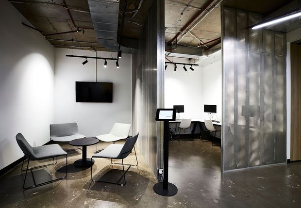 An open-plan contemporary commercial interior with a casual lounge area in the foreground and shared workstations behind it. The space features white walls, polished concrete floors, exposed services and ceiling structure, black track lighting, a wall-mounted screen, metal-clad columns, and translucent corrugated partition panels.