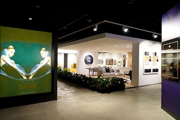 A contemporary interior showroom or design exhibition space with polished concrete flooring, a black exposed ceiling, track lighting, white display walls, framed artwork and material samples, and a staged living area beyond a low indoor planting bed. A large illuminated artwork panel and branded display feature the entrance zone.