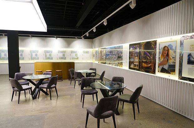 A contemporary gallery-like commercial interior with multiple small café-style tables and upholstered chairs arranged on a polished concrete floor. The space features ribbed white wall panelling, illuminated display panels and framed imagery along the walls, a dark exposed ceiling with track lighting, a curved mustard-coloured counter, and a large geometric ceiling light.