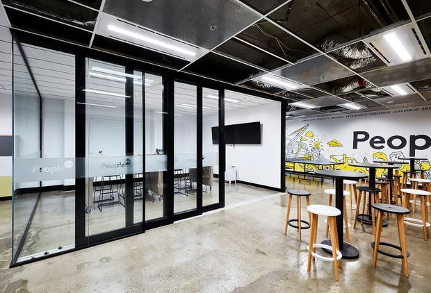 A contemporary commercial office and collaborative breakout area with black-framed glass meeting rooms, polished concrete flooring, exposed services and an acoustic ceiling grid. A large illustrated mural and high communal tables with stools create an informal, creative workspace atmosphere.