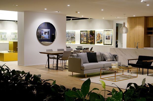 A refined open-plan interior combining a living room, dining area, and kitchen. The space features a low contemporary ceiling with recessed lighting, pale timber flooring, a neutral sofa arrangement, a slim glass coffee table with a brass-toned frame, a dark dining table with upholstered chairs, a marble-look kitchen island, and warm timber cabinetry. A round wall mirror and framed artwork add visual interest, while indoor greenery occupies the foreground.