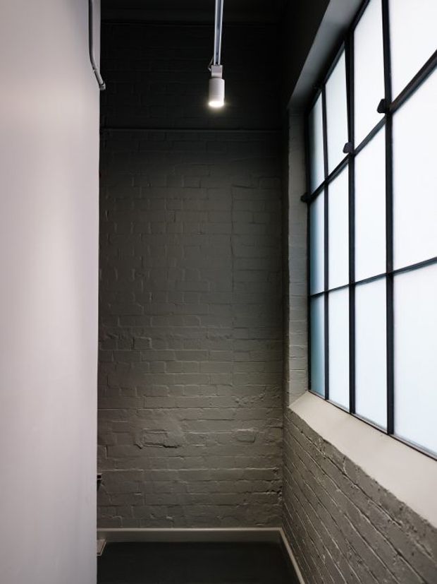 A narrow, tall interior space with exposed painted brick walls, a dark painted ceiling, black-framed industrial-style grid windows, and a single suspended pendant light. The minimal, unfinished material palette creates a strong loft-like architectural character.