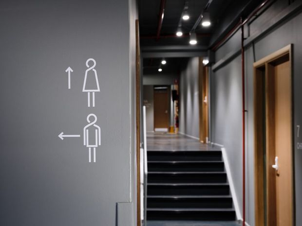 A modern interior hallway with a directional restroom sign on a grey wall, dark stair treads leading to an upper corridor, exposed ceiling services, recessed and track lighting, and timber-framed doors. The space has an industrial, functional character with a monochromatic grey, black, and timber palette.