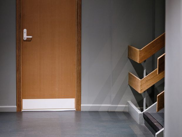 A compact interior hallway or stair landing with a plain timber door, grey walls, polished grey floor, and a staircase with angular timber handrails and dark steps. The scene has a clean, functional, modernist character with strong geometric lines and contrasting wood and grey finishes.