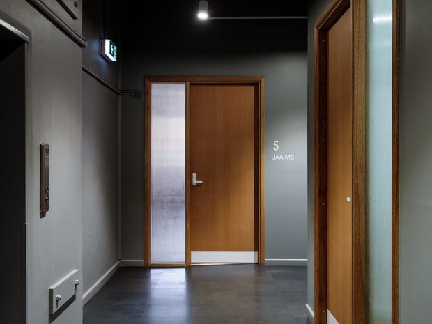 A dark, refined commercial or apartment-building hallway with charcoal painted walls, polished dark flooring, timber-framed doors, and frosted glass panels. A central timber door is marked “5 JAAIMS,” with additional glazed doorways visible at the right and an illuminated exit sign overhead.