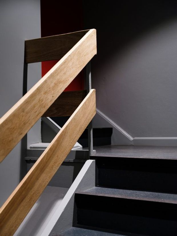 Close-up architectural photograph of a modern stairway with black stair treads and risers, grey walls, and angular light timber handrails supported by dark metal framing. A red accent wall is visible through the stair structure.