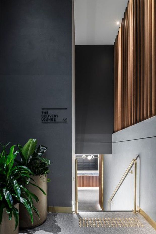 A dramatic, refined interior stairway descending through a tall modern space. Dark charcoal walls and grey textured flooring contrast with warm vertical timber battens on the upper wall. Brass or gold-toned handrails, recessed lighting, a patterned metal stair tread, and a large leafy planter create a sophisticated hospitality or commercial entry atmosphere. Signage reads “The Delivery Lounge.”