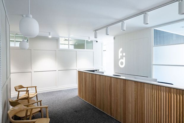 Bright, minimalist commercial reception area with a curved front desk clad in vertical timber slats, white paneled walls, a grey carpeted floor, contemporary pendant and track lighting, and timber-framed lounge chairs. Large windows and a white branded wall feature add to the clean, professional setting.