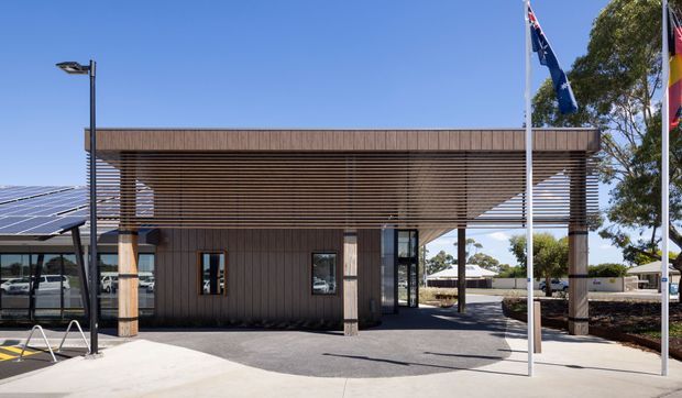 Modern single-storey civic or commercial building entrance with a prominent timber-look horizontal louvre screen and substantial square columns. Glazed doors and windows open onto a paved forecourt, with solar panels visible on the adjoining roof and flagpoles in front. The composition is clean and contemporary, with strong linear shading elements and a bright blue-sky setting.